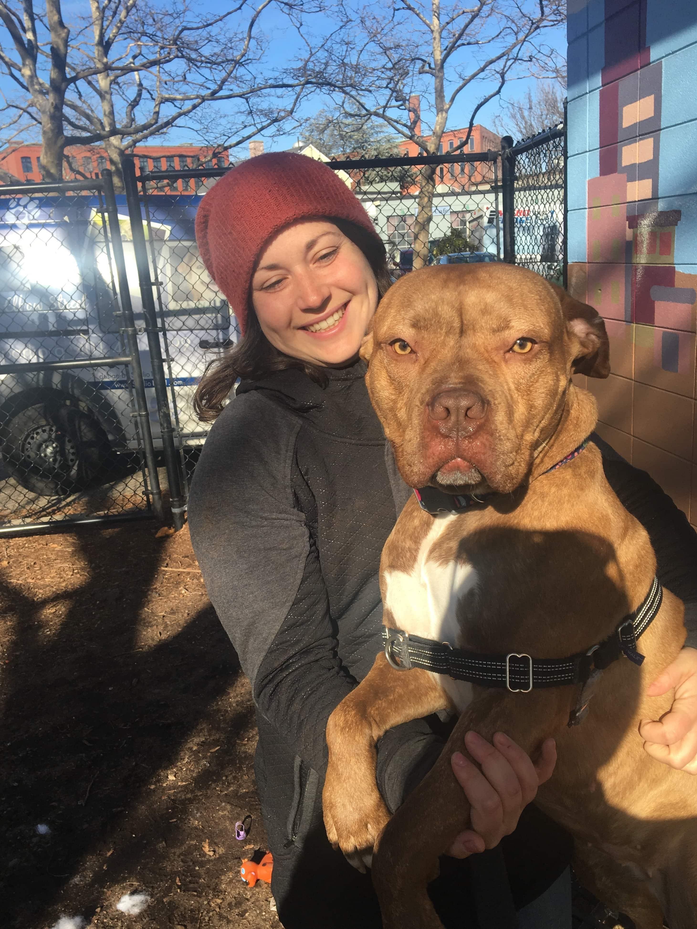 Woman in red hat holds brown shelter dog outdoors.